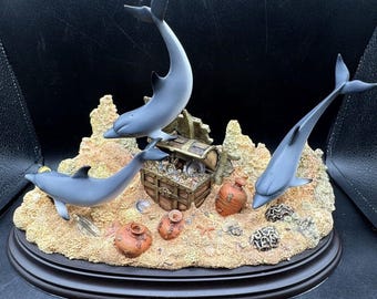 May include: A figurine of three dolphins jumping out of the water, surrounding a treasure chest filled with gold coins. The dolphins are gray and white, and the treasure chest is brown with gold accents. The figurine is set on a brown base with a sandy texture.