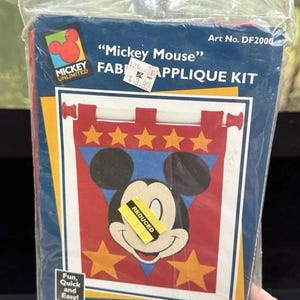 May include: A Mickey Mouse fabric applique kit with a red, white and blue banner featuring Mickey Mouse's face and five yellow stars. The kit includes instructions and all the materials needed to create the banner. Art No. DF2000.