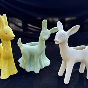 Puede incluir: Tres figuritas de ciervos de cerámica, una amarilla, una verde y una blanca, están dispuestas sobre una superficie negra. Cada ciervo tiene una pequeña maceta en la espalda.