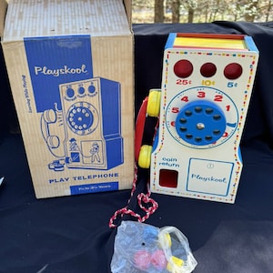 May include: Vintage Playskool toy telephone with a red, yellow, and blue rotary dial. The phone has a coin return slot and a Playskool logo. The box is brown with a blue and white illustration of the phone.
