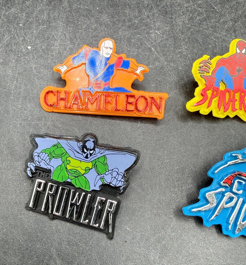 Może przedstawiać: Kolekcja kolorowych przypinek z postaciami superbohater&oacute;w w stylu vintage. Przypinki przedstawiają postacie takie jak Chameleon, Spider-Man i The Prowler, z odważnymi kolorami i tekstem. Przypinki wykonane są z błyszczącego materiału.
