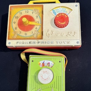 Puede incluir: Dos radios de juguete Fisher-Price vintage. La radio superior es de color blanquecino con un asa amarilla y detalles en rojo. Cuenta con una esfera de reloj y un dial que dice "Hickory Dickory Turn". La radio inferior es verde con un dial blanco y una correa a rayas.