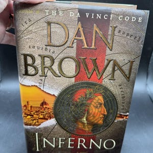 May include: A hardcover book titled Inferno by Dan Brown, author of The Da Vinci Code. The cover features a portrait of Dante Alighieri overlaid with a circular map design, a red vertical stripe, and a cityscape of Florence at the bottom left.