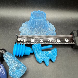 May include: Several translucent blue plastic toy parts are displayed against a dark background. A ruler shows measurements in inches. The parts include a water-like base, a cylindrical piece, and a textured, stylized shape.
