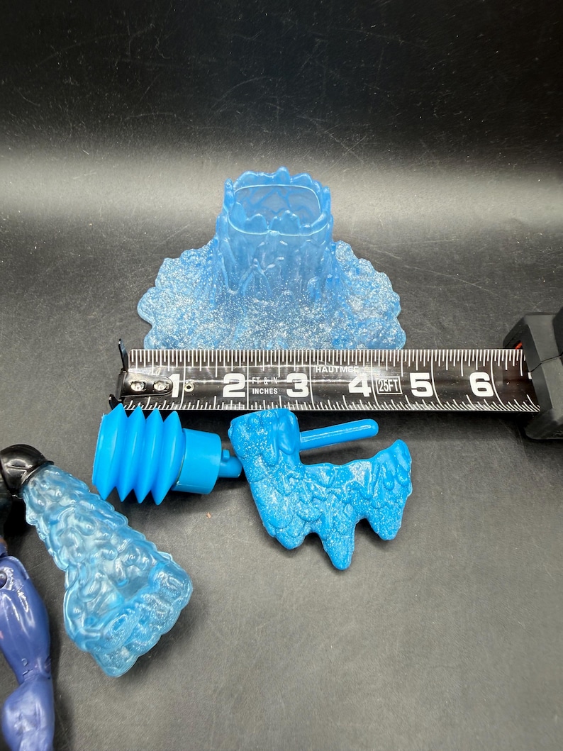 May include: Several translucent blue plastic toy parts are displayed against a dark background. A ruler shows measurements in inches. The parts include a water-like base, a cylindrical piece, and a textured, stylized shape.