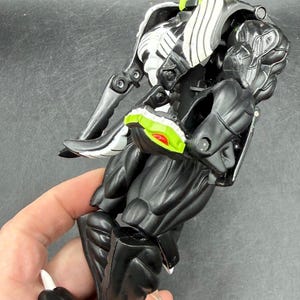 May include: A black and white action figure of a snake-like creature with a red tongue and green accents. The figure is held in a hand and has articulated joints. The toy is made of plastic.