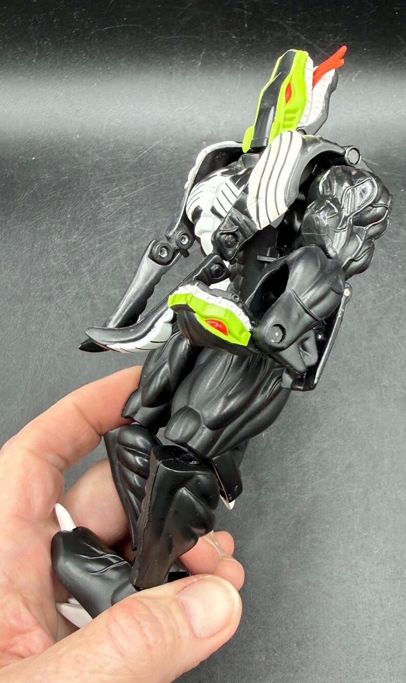 May include: A black and white action figure of a snake-like creature with a red tongue and green accents. The figure is held in a hand and has articulated joints. The toy is made of plastic.