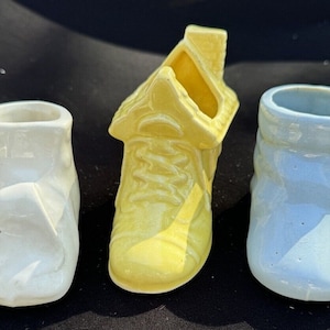 以下が含まれることがあります： 異なる色のセラミック製のベビーシューズ3足。靴は白、黄、水色です。すべて伝統的なベビーシューズの形をしています。