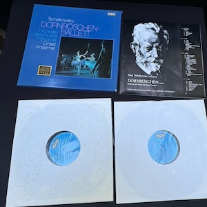 Puede incluir: Una colección de discos de vinilo vintage con música clásica. Las portadas de los álbumes son azules y negras, con texto en varios idiomas. Los discos están en fundas blancas con etiquetas azules. El logotipo de Decca es visible.