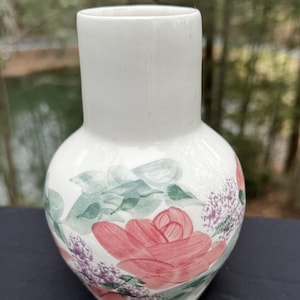 May include: A white ceramic vase with a floral design. The vase has a round body and a narrow neck. The flowers are painted in shades of pink, green, and purple.