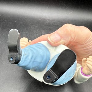 May include: A ceramic figurine of a person lying down, wearing a white coat, light blue pants, and black shoes. The figurine's hands are clenched into fists. The figurine is being held by a person.