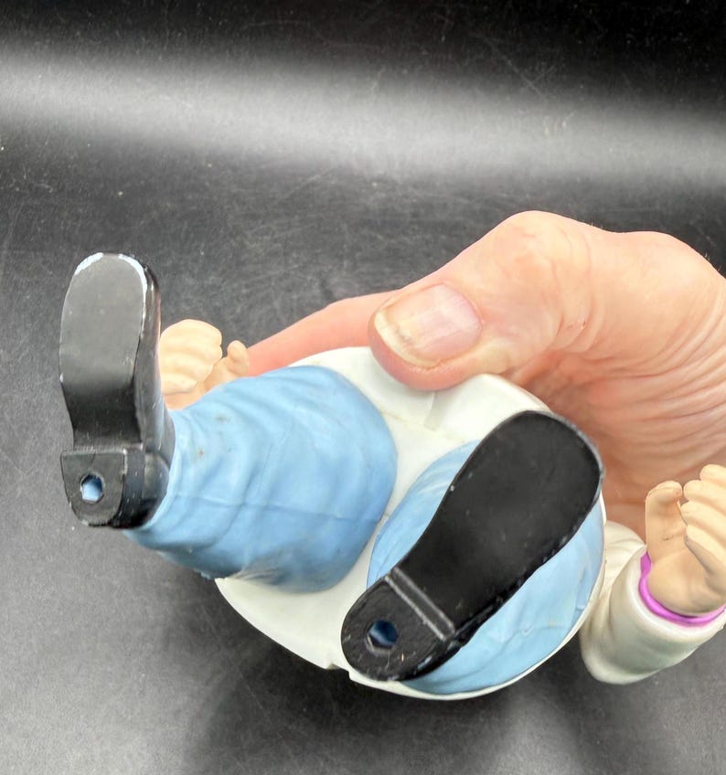 May include: A ceramic figurine of a person lying down, wearing a white coat, light blue pants, and black shoes. The figurine's hands are clenched into fists. The figurine is being held by a person.