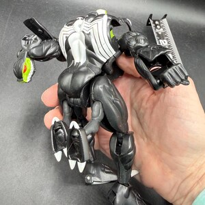 May include: Black and white action figure of a Venom-like character with articulated joints. The figure has a white spider-like design on its back and head, with green accents on the eyes and mouth. The figure is approximately 15 cm tall.