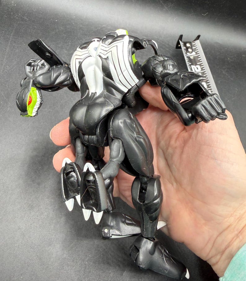 May include: Black and white action figure of a Venom-like character with articulated joints. The figure has a white spider-like design on its back and head, with green accents on the eyes and mouth. The figure is approximately 15 cm tall.