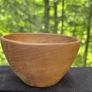 Puede incluir: Un pequeño cuenco de madera hecho a mano con un acabado marrón claro natural. El cuenco tiene un diseño liso y curvado y una textura de veta de madera visible. El cuenco probablemente se utiliza para servir o exhibir.