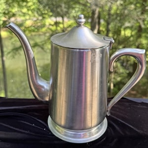 Puede incluir: Una cafetera de metal plateado con un pico y asa curvados. La cafetera tiene una tapa con un pequeño pomo decorativo en la parte superior. La cafetera se asienta sobre una base redonda.