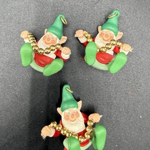May include: Three ceramic Christmas elf ornaments. Each elf wears a green hat, red jacket, and green shoes. They hold a gold bead garland. The elves have rosy cheeks and white beards. Each ornament has a gold loop for hanging.