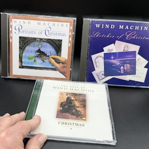 May include: Three Wind Machine Christmas music CDs in clear plastic jewel cases. The top left cover is tan with a hand painting on sheet music. The top right cover is blue with sketches. The bottom cover is white with a central photo.