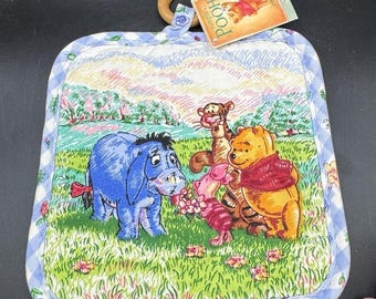 May include: A Winnie the Pooh themed pot holder with a wooden handle. The pot holder features a colorful illustration of Pooh, Tigger, Piglet, and Eeyore in a grassy field. The border is a blue and white gingham pattern.