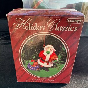 Puede incluir: Una caja roja y blanca con un adorno de Navidad de Santa Claus en su interior. El adorno es parte de una serie llamada "It Must Be St. Nick" y se titula "Checking It Twice". La caja está etiquetada como "Holiday Classics" y "Eckerd".