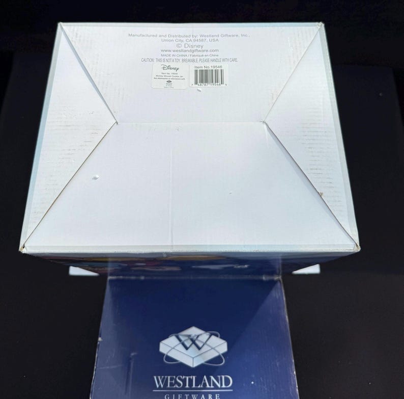 May include: A white cardboard box with a folded top, featuring text from Westland Giftware and Disney. The box is on top of a dark blue box with the Westland Giftware logo. The box has a warning label.