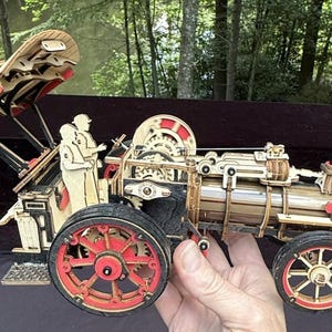 May include: A mechanical model of a steam traction engine constructed from laser-cut wood pieces. The model features red wheels, a black smokestack, exposed gears, and two small wooden figures seated in the rear cab area under a hinged roof.