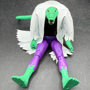 K&ouml;nnte beinhalten: Eine gr&uuml;ne, echsenartige Actionfigur mit einem wei&szlig;en Laborkittel, lila Hose und gr&uuml;nen Stiefeln. Die Figur hat einen langen gr&uuml;nen Schwanz und einen detaillierten Reptilienkopf. Ein Sammlerspielzeug.