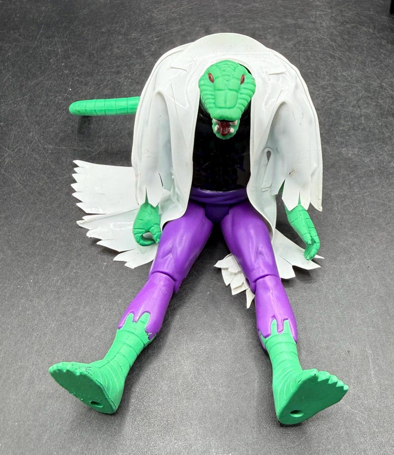 K&ouml;nnte beinhalten: Eine gr&uuml;ne, echsenartige Actionfigur mit einem wei&szlig;en Laborkittel, lila Hose und gr&uuml;nen Stiefeln. Die Figur hat einen langen gr&uuml;nen Schwanz und einen detaillierten Reptilienkopf. Ein Sammlerspielzeug.