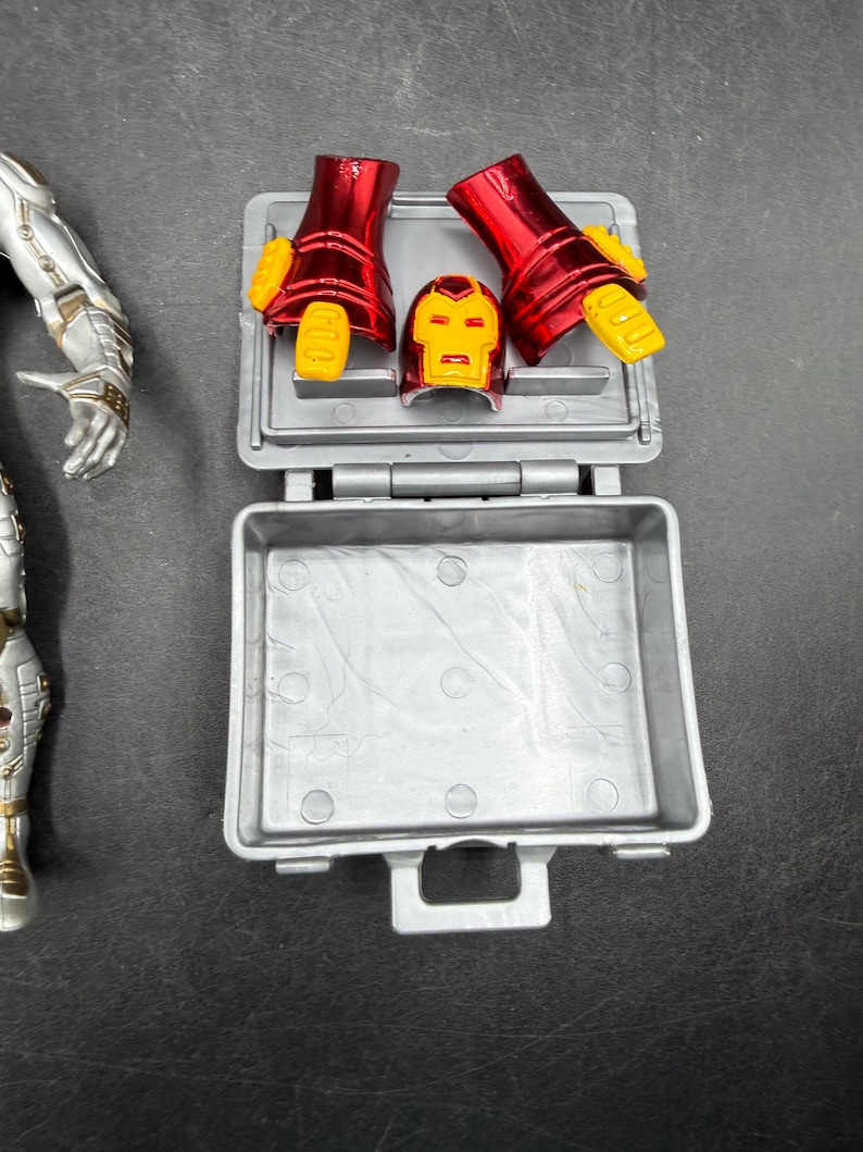 May include: A silver and gold action figure with red and yellow Iron Man armor pieces. The armor pieces include gauntlets, a helmet, and a gray plastic case.