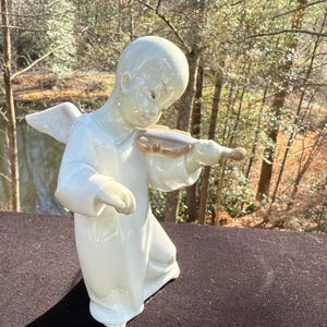Pode incluir: Estatueta de anjo de cerâmica branca tocando violino. O anjo é retratado com asas e uma túnica fluida. O violino é de cor castanho claro. A estatueta está situada em um cenário de árvores e um corpo de água.