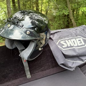 Pode incluir: Um capacete de motocicleta Shoei preto brilhante com viseira e alça de pescoço cinza. O capacete está ao lado de uma bolsa de capacete Shoei cinza. O logotipo da Shoei é visível no capacete e na bolsa.
