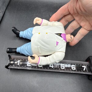 May include: A plastic action figure of a character in a white suit, blue pants, and black shoes. The figure is approximately 6 inches long. The figure has a bald head and is lying on a black measuring tape.