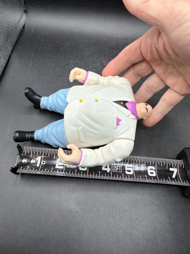 May include: A plastic action figure of a character in a white suit, blue pants, and black shoes. The figure is approximately 6 inches long. The figure has a bald head and is lying on a black measuring tape.