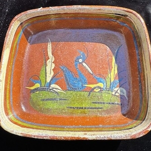 May include: A square terracotta dish with a hand-painted design of two blue birds in a green pond with yellow and white reeds. The dish has a brown glaze with a yellow and white border.