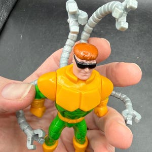 Puede incluir: Una figura de acci&oacute;n del Dr. Octopus con un traje amarillo y verde, pelo naranja y gafas de sol negras. La figura tiene cuatro brazos mec&aacute;nicos grises articulados. El juguete se sostiene en una mano.