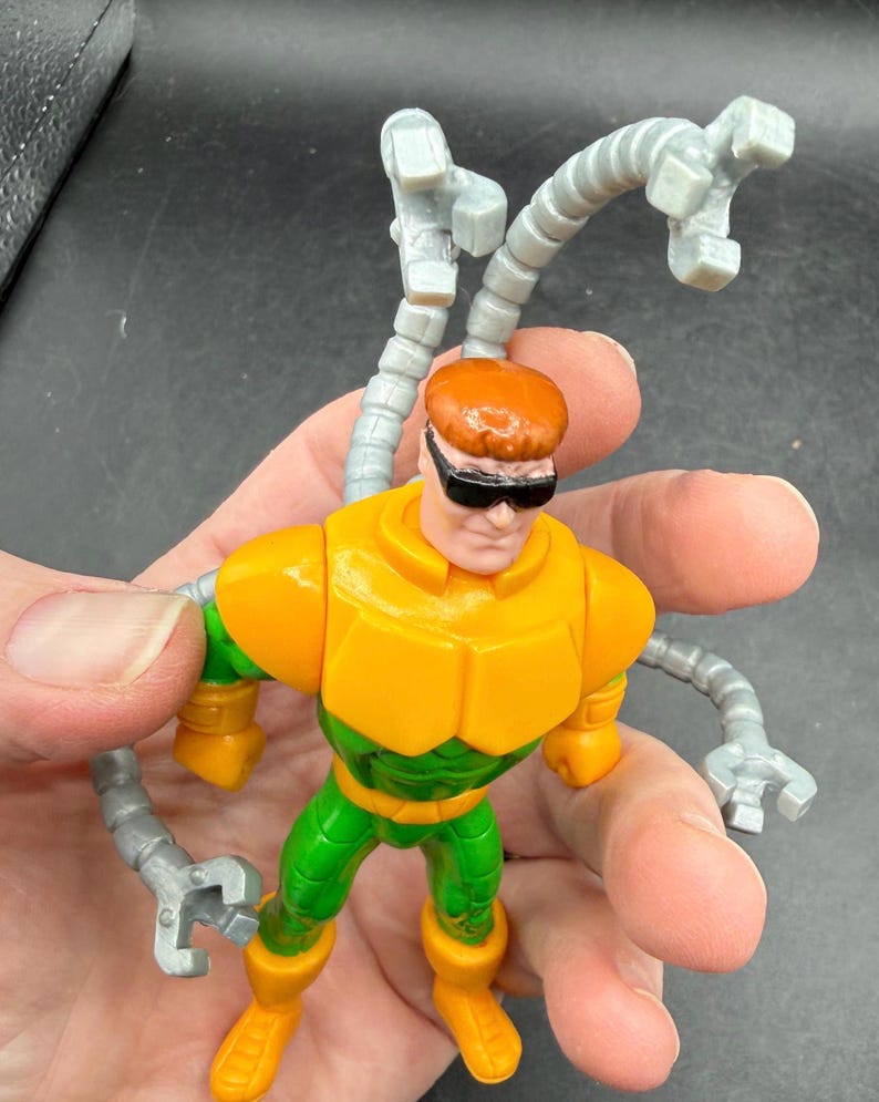 Puede incluir: Una figura de acci&oacute;n del Dr. Octopus con un traje amarillo y verde, pelo naranja y gafas de sol negras. La figura tiene cuatro brazos mec&aacute;nicos grises articulados. El juguete se sostiene en una mano.