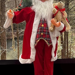 Pode incluir: Uma figura decorativa do Pai Natal, vestida com um fato de veludo vermelho com acabamentos brancos. Ele segura um ursinho de pelúcia e uma pequena árvore de Natal. A figura tem barba branca, óculos e uma expressão alegre, ideal para a decoração festiva.