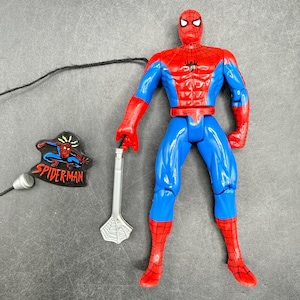 May include: A Spider-Man action figure in a classic red and blue suit, holding a silver web shooter. A Spider-Man patch and an earbud are also present. The figure has a detailed design, capturing the iconic superhero's appearance.
