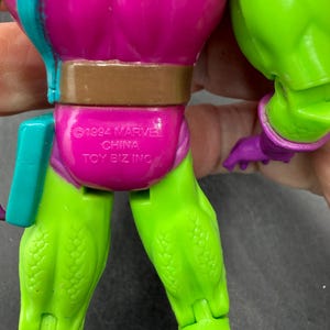 May include: A close-up of a vintage action figure. The figure has a purple torso, green legs, and a brown belt. The text "&copy;1994 MARVEL CHINA TOY BIZ INC" is printed on the figure's back.