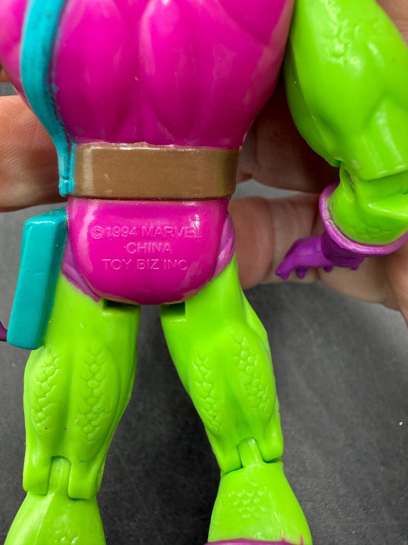 May include: A close-up of a vintage action figure. The figure has a purple torso, green legs, and a brown belt. The text "&copy;1994 MARVEL CHINA TOY BIZ INC" is printed on the figure's back.