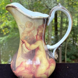 May include: A decorative ceramic pitcher with a classical painting of cherubs. The pitcher has a white handle with gold trim, a light blue rim, and a beige body. The artwork features cherubs in shades of yellow, pink, and red.