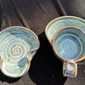 May include: Two handmade ceramic bowls with a blue and green glaze. The bowls have a swirl pattern and a handle.