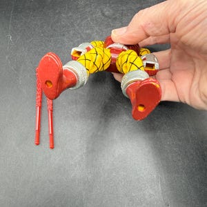 以下が含まれることがあります： 関節が可動する赤、黄、銀色のフィギュア。フィギュアは赤いブーツ、黄色のセグメント化された手足、赤い胴体を持っています。2つの赤いアクセサリーが含まれています。フィギュアは暗い背景に対して保持されています。