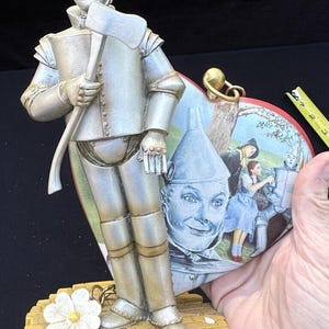 May include: A silver figurine of the Tin Man from The Wizard of Oz holding an axe. The figurine is standing on a yellow brick road base with a white flower. The Tin Man is holding a heart-shaped picture frame with a scene from the movie.