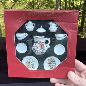 Puede incluir: Juego de té de porcelana en miniatura en una caja roja. El juego incluye una tetera, una jarra de crema, un azucarero, dos tazas, dos platillos y dos platos. La tetera y los platos presentan ilustraciones coloridas. La caja dice "Mini M.J. Hummel Set".