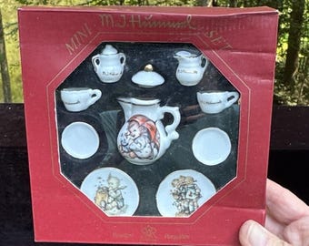 Op de afbeelding: Miniatuur porseleinen theeset in een rode doos. De set bevat een theepot, melkkan, suikerpot, twee kopjes, twee schoteltjes en twee borden. De theepot en borden hebben kleurrijke illustraties. Op de doos staat "Mini M.J. Hummel Set".