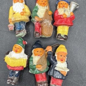 Op de afbeelding: Een verzameling van zes kleine, kleurrijke kabouterbeeldjes. Elke kabouter heeft een witte baard, roze wangen en een puntmuts. Ze zijn gekleed in verschillende outfits, sommige met muziekinstrumenten, andere met voorwerpen. De beeldjes zijn in heldere kleuren geschilderd.