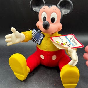 May include: A vintage Mickey Mouse doll, featuring a yellow shirt, red pants, and yellow shoes. The doll has black ears, a pink face, and white gloves. It has a tag that reads "Disney PLAYMATES by applause."