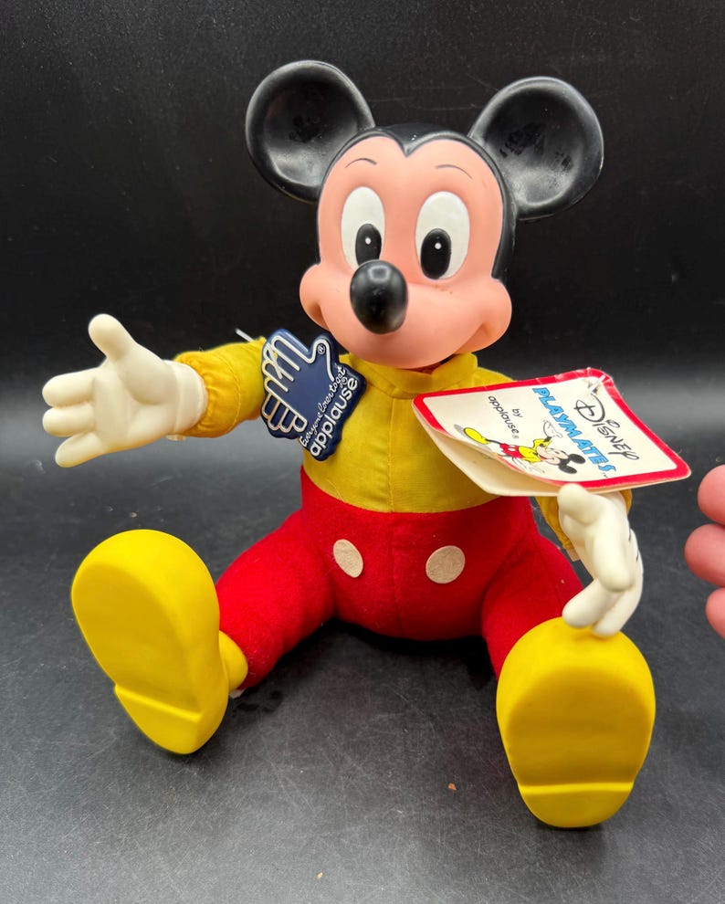 May include: A vintage Mickey Mouse doll, featuring a yellow shirt, red pants, and yellow shoes. The doll has black ears, a pink face, and white gloves. It has a tag that reads "Disney PLAYMATES by applause."