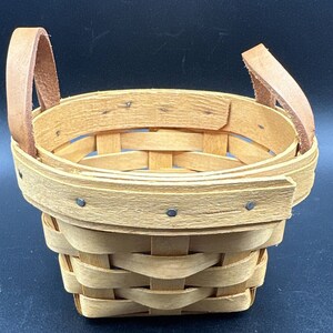 May include: A small, woven basket with two leather handles. The basket is made of natural wood and has a light brown colour.
